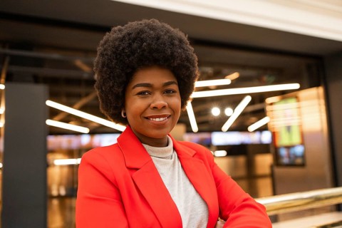 Confident woman in a vibrant red blazer stands smiling indoors. Modern lighting and a blurred