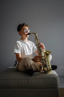A boy in glasses sits cross legged on a cushion, playing a saxophone. Wearing a white T shirt, he