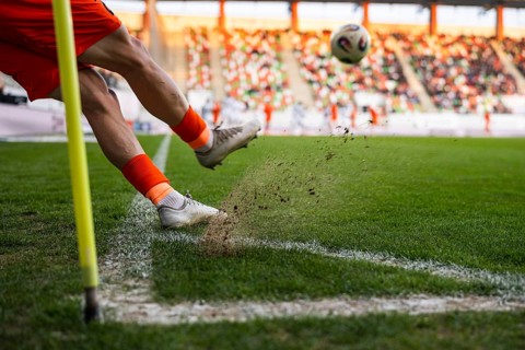 A close-up of a soccer player's legs kicking a ball during a corner kick with turf and dust flying