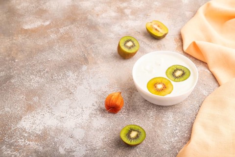 Yogurt with kiwi in white bowl on brown concrete background and orange linen textile, side view,