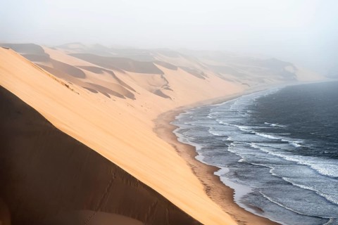 Sand dunes with wave patterns and sea in evening light, wind blowing sand across the dunes,