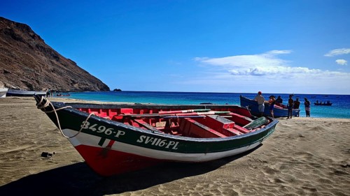 Two colorful boats are lying on the beach, with several people standing around a boat, hiking, Sao