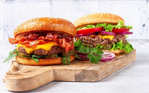 Two burgers with beef patty, fried bacon, cheddar cheese, tomatoes, on a light table
