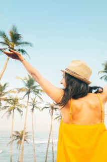 Carefree happy woman traveler in yellow summer dress and straw hat enjoy palm beach in tropical