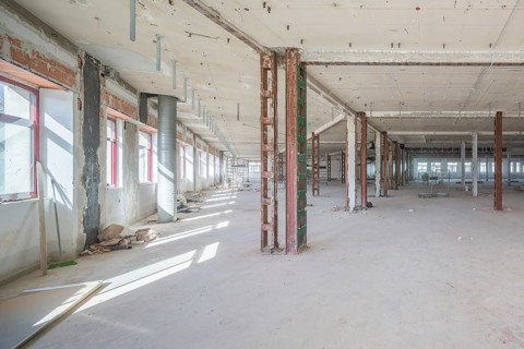 A large, empty industrial space with exposed beams and scattered construction materials. Sunlight