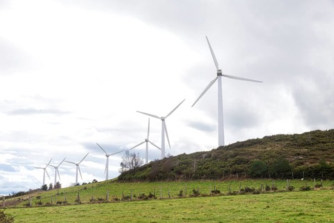 A row of windmills lines a lush, green hillside, harnessing wind energy beneath a cloudy sky. The