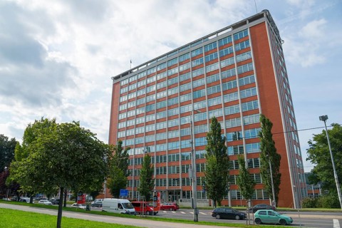 Large brick building in an urban environment with passing cars and trees on a cloudy day, Batuv
