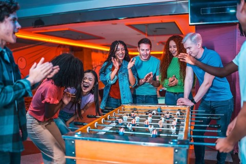 Multiethnic friends enjoying a foosball game, clapping and laughing, inside a vibrant entertainment