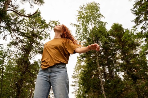 Woman in a mustard shirt and jeans, joyfully embracing nature in a lush Swedish forest. Surrounded