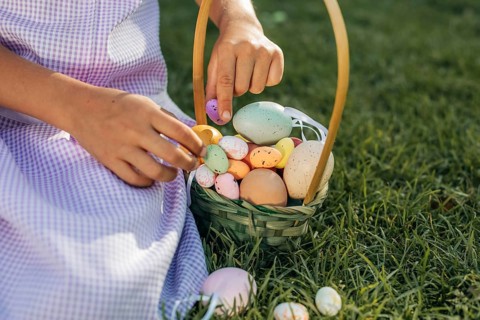 A child in a purple dress gathers pastel colored Easter eggs into a woven basket, resting on