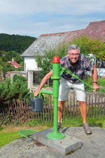 A smiling elderly gentleman fills a watering can at a green water pump in a rural setting, Lipnice