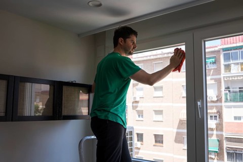 A man performs household chores in an urban apartment, focusing on cleaning the window. The image