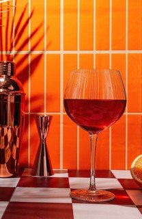 A cocktail glass with a red drink stands on a checkered table. Nearby, a cocktail shaker and a