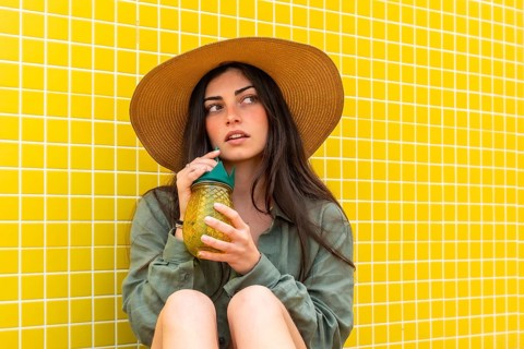 Woman wearing a straw hat and green shirt, taking a sip from a pineapple shaped jar while sitting