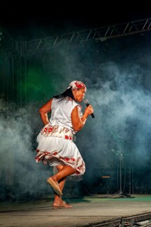 Woman dances and sings vividly on a smoky stage in traditional dress, a band plays and dances at a