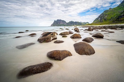 Rocks on Uttakleiv sandy beach, coast and blue sea, long exposure, rocky mountain peaks,
