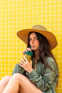 Young woman sitting by a bright yellow tiled wall, enjoying a cool drink from a pineapple shaped