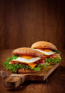 Two juicy burgers sit on a wooden board, each filled with lettuce, bacon, cheese, and fried eggs