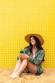 Young woman sits on warm sand against a yellow tiled wall, smiling and holding a pineapple shaped