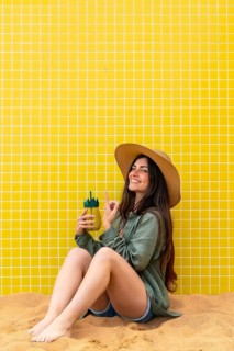 Happy young woman sitting in sand against a yellow tiled wall, holding a refreshing pineapple