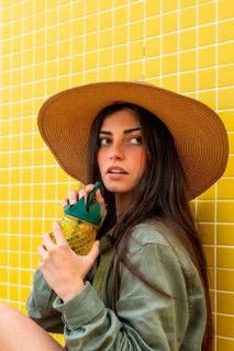 Young woman wearing a straw hat and green shirt, sitting against a bright yellow tiled wall,