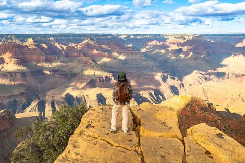 Person wearing a cap and backpack standing on a rock viewpoint, admiring the vast layers of rock