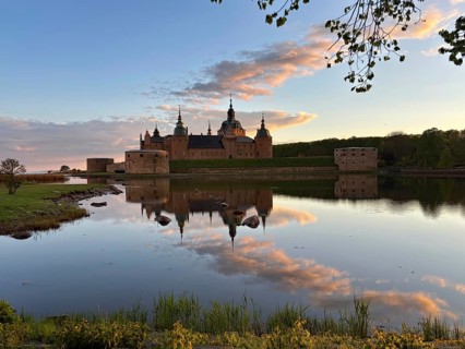 A castle is reflected in a calm lake, surrounded by colorful evening skies and peaceful nature,