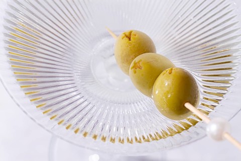 A clear glass cocktail sits on a white marble surface. Three green olives are placed on a skewer