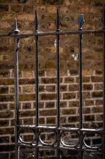 Old black painted metal fence with cobwebs in front of a weathered brick wall, Chelsea, London,