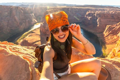 Young woman smiling and adjusting sunglasses, sitting on sandstone cliffs overlooking the colorado