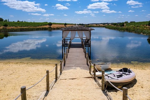 A wooden walkway leads to a pavilion over a tranquil lake surrounded by nature, Pedrógão, Beja,