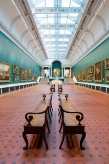 An elongated gallery room with benches, skylight and numerous historic paintings on green walls,