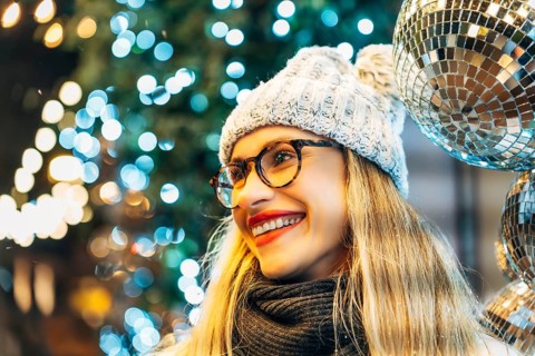 Young woman in winter hat in front of festive lights. A woman wearing glasses, a gray knit winter