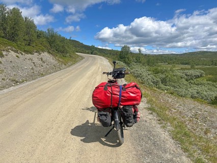 A bicycle with red bags is parked on a dirt road in the middle of green countryside, Peer Gynt