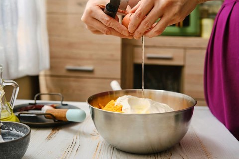 A person is cracking an egg into a metal bowl filled with yellow ingredients. The kitchen has