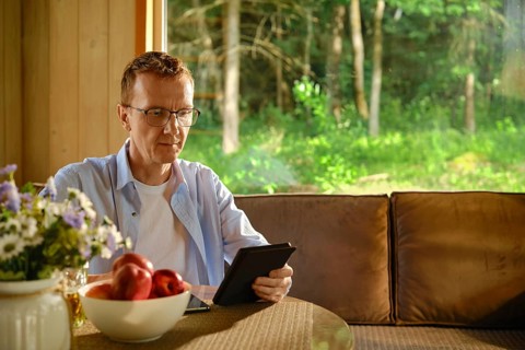 Middle-aged man in glasses sits at table in wooden cabin with e-book. Sunlight enters room through