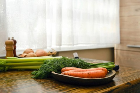 Vegetables are on a wooden table in a kitchen. Carrots, dill and celery are arranged neatly on a