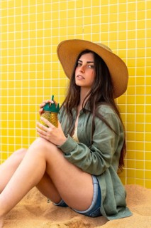Woman wearing straw hat and linen shirt sitting on sand against yellow tiled wall, holding a