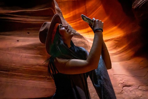 Young woman in a cowboy hat smiling, holding a smartphone up to capture the undulating sandstone