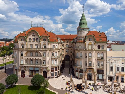 Large baroque building with red roof and green tower in a busy square under cloudy sky, aerial