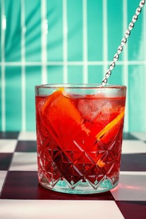 A cocktail with orange slices rests in a glass on a black and white checkered table. The background