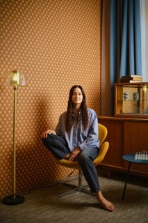 Woman with long hair sits in yellow chair at home. Wall has unique pattern. Floor lamp stands