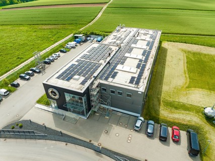 Building with solar panels on the roof, surrounded by fields and parked cars, PV assembly, Perrot