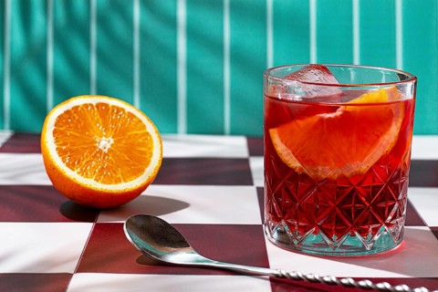 A clear glass holds a colorful drink with slices of orange. Next to it, there is a halved orange on