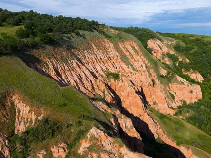 Hilly rocky landscape with traces of erosion, surrounded by green trees and a blue sky, aerial