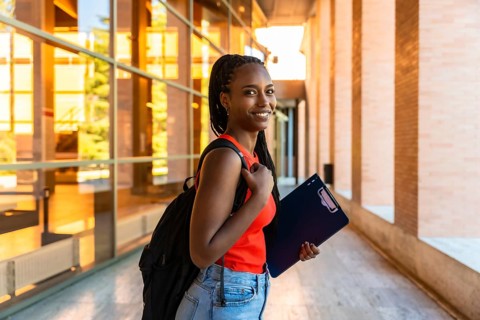Confident young black woman student with backpack and folder smiling, walking on a modern