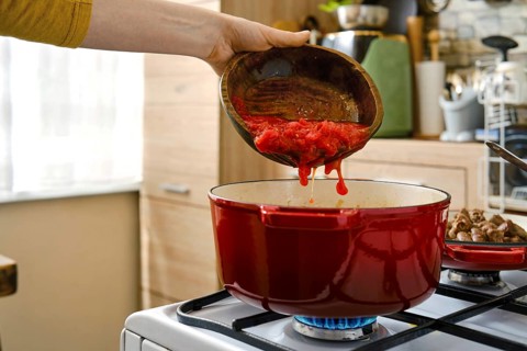 A person pours chopped tomatoes from a bowl into a red pot on a stove. The kitchen has wooden