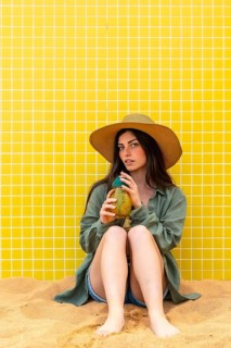 Young woman sitting on the sand, wearing a straw hat and casual clothes, holding a pineapple shaped