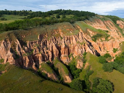 Impressive erosion formations in earthy hills surrounded by lush vegetation under blue sky, aerial
