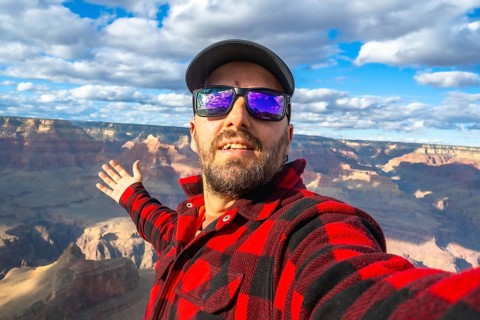 Bearded man wearing sunglasses, a cap, and a plaid shirt, smiling and gesturing towards the vast